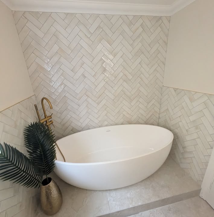 Freestanding oval bath with a brushed-gold tap, set against cream herringbone wall tiles